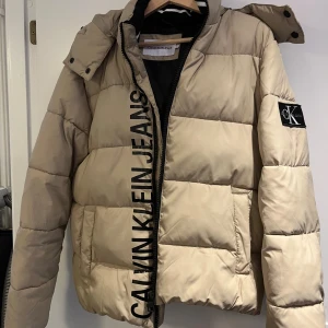Beige pufferjacka från Calvin Klein Jeans - Säljer en beige pufferjacka från Calvin Klein Jeans med stor svart logga längs dragkedjan och patch på ärmen. Jackan har huva, svarta detaljer och är quiltad för extra värme. Perfekt för kalla dagar och har en clean, modern look. Jackan är som ny använd max 2 gånger. Utgångspris 1500