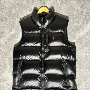 Svart dunväst från Polo Ralph Lauren - Snygg svart dunväst från Polo Ralph Lauren med röd broderad logga på bröstet. Västen har hög krage, tre dragkedjefickor och quiltad design. Perfekt för lager-på-lager och har en stilren look med glansigt material.