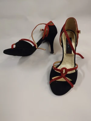 Svarta och röda Dansskor Nueva epoka - Snygga Danskor med klack från Nurva epoka i svart  med röda och guldfärgade remmar. Skorna har öppen tå, rund tå och elegant spänne vid ankeln. Insidan är guldfärgad och ger en lyxig känsla. Perfekt för dig som gillar att sticka ut med stilrena detaljer. Nypris 1500-1800kr