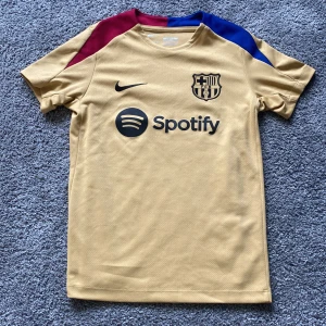 Barcelona guld fotbollströja Nike M - Säljer en Barcelona fotbollströja i guld från Nike, slim fit och tillverkad i Dri-Fit material. Tröjan har korta ärmar, klubbmärke på bröstet och Spotify-logga. Snygga detaljer i rött och blått på axlarna. Perfekt för dig som älskar Barca!