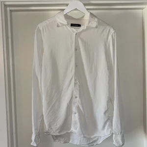 Vit skjorta från J.Lindeberg, stl M - Klassisk vit skjorta från J.Lindeberg i storlek M. Skjortan har lång ärm, knappar framtill och diskret broderad logga på framsidan. Tillverkad i mjuk bomull med stilren krage och enkel design som funkar till många olika looks. Köpte ny för 1400kr.