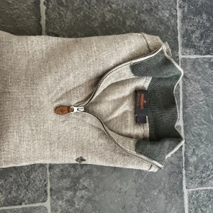 Beige half zip tröja Morris - Snygg beige långärmad tröja från Morris i 100% merinoull. Tröjan har en half zip med läderdetalj på dragkedjan och ribbade muddar i mörkgrått vid krage, ärmslut och nederkant. Perfekt för dig som gillar stilrena och klassiska plagg.