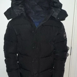 Svart dunjacka från Canada Goose - Säljer en svart dunjacka från Canada Goose med huva och snyggt broderat märke på ärmen. Jackan har dragkedja och knappar framtill, quiltad design och långärmad modell. Perfekt för kalla vinterdagar och riktigt stilren look. helt ny oandvänd min vän har samma annons ute hjäper honom.