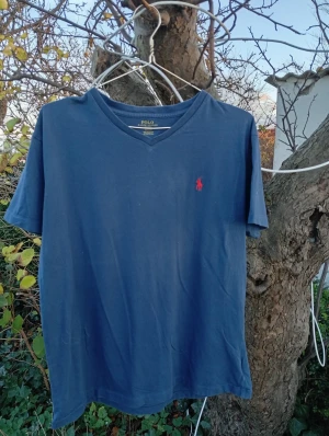 Blå t-shirt från Polo Ralph Lauren - Snygg blå t-shirt från Polo Ralph Lauren med klassisk v-ringning och röd broderad logga på bröstet. T-shirten är kortärmad och tillverkad i mjuk bomull, perfekt för en avslappnad och stilren look.