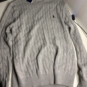 Grå kabelstickad tröja Polo Ralph Lauren - Supersnygg grå kabelstickad tröja från Polo Ralph Lauren med klassisk rund hals och broderad logga på bröstet. Tröjan har långa ärmar och ribbade muddar vid ärmslut och nederkant. Perfekt för dig som gillar stilren och tidlös design.