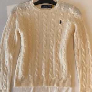 Vit kabelstickad tröja Polo Ralph Lauren - Kabelstickad tröja i vit från Polo Ralph Lauren med klassisk rund hals och broderad marinblå logga på bröstet. Tröjan har långärmad passform och ribbade muddar vid ärmslut och nederkant. Perfekt för dig som gillar stilren och tidlös design. Endast använd en gång