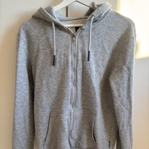 Grå hoodie från Only & Sons - Säljer en grå hoodie från Only & Sons med dragkedja framtill, huva med snörning och två fickor. Mjuk insida i fleece och klassisk passform. Perfekt för chill dagar och enkel att matcha med jeans eller joggers.