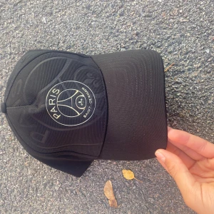 Svart PSG keps med guldtryck - Snygg svart keps från Paris Saint Germain med guldtryckt logga framtill och texten 'ICI C'EST PARIS' broderad baktill. Justerbar rem med PSG-detalj. Perfekt för dig som älskar fotboll och vill ha en cool accessoar.