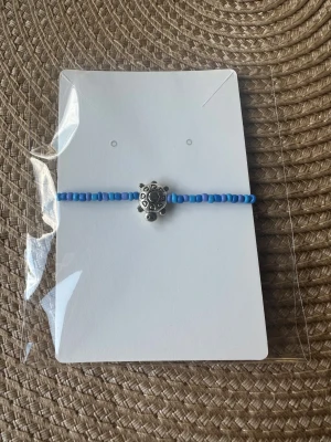 Blått pärlarmband med sköldpadda - Snyggt hemmagjort armband med små blå pärlor och en silverfärgad sköldpadda i mitten. Perfekt accessoar för dig som gillar djurmotiv och vill ha något unikt på handleden. Armbandet är enkelt och stilrent, passar till många olika looks.