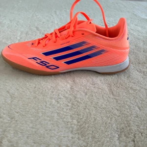 Adidas F50 orange fotbollssko - Säljer  Adidas F50 fotbollsskor i en riktigt snygg orange färg med blå detaljer och logga på sidan. Skorna har snörning, platt gummisula och är designad för inomhusspel. Materialet är syntet och skorna har en spetsig tå för bättre bollkontroll. Om det är otydligt är det 2 skor.