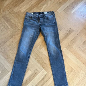 Grå slim fit jeans från Jack & Jones - Säljer ett par grå slim fit jeans från Jack & Jones med snygg tvättad look och klassiska fem fickor. Jeansen har knappgylf, bälteshällor och broderade bakfickor. Perfekta för dig som gillar en smalare passform och stilren design. Hör av er vid frågor