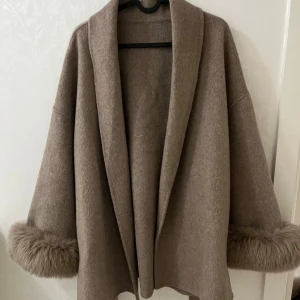 Beige kappa med pälsdetalj - Säljer en beige kappa med bred krage och fluffiga pälsdetaljer vid ärmsluten. Jackan har en loose passform och är tillverkad i mjukt ullmaterial. Helt ny