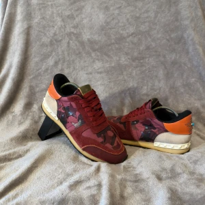 Valentino Rockrunner sneakers röd/orange - Snygga Valentino Rockrunner sneakers med röd och orange färg, camouflagemönster och stjärndetaljer med nitar och turkos sten på hälen. Skorna har snörning, platt sula och är tillverkade i mocka, skinn och syntet. Ikoniska pyramidnitar på bakkappan ger extra edge. Storlek 41 men skulle säga att de passar 41,5. Skick 9/10