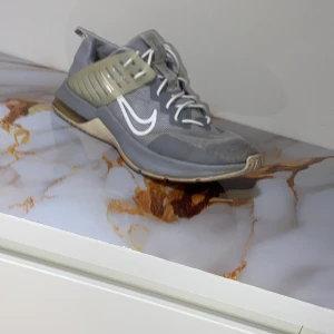 Grå Nike sneakers med vit swoosh - Snygga grå Nike sneakers med vit swoosh på sidan och ljusgrå detaljer. Skorna har snörning och en robust platt sula med transparent sektion vid hälen. Ovandelen är i syntetmaterial och mesh för bra ventilation. Perfekt för dig som gillar sportig stil. Men är lite taita så skulle säga de också passar storlek 44-45.