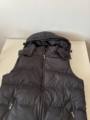 Svart dunväst med huva och dragkedja - Svart dunväst med avtagbar huva och två fickor med dragkedja. Västen har quiltad design och hel dragkedja framtill. Perfekt för lager-på-lager och chill höstdagar. Materialet är syntet och dun, vilket gör den både lätt och varm. Helt ny.