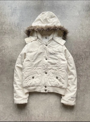 Vit 2000s y2k pufferjacka  - Snygg vit vinterjacka från College Athlete med huva och fuskpäls. Jackan har broderad logga på bröstet, knappar framtill och två fickor med lock. Ribbad nederkant och ärmslut ger en schysst passform. Mått: 51x45 cm. Strl: XXS / 152