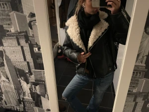 Svart pälskantad skinnjacka - Säljer en svart skinnjacka med stor krage i beige fuskpäls. Jackan har dragkedja framtill, detaljer med spännen vid midjan och ärmarna, samt en loose passform. Perfekt för dig som gillar en cool och trendig look med extra värme runt halsen. Väldigt varm så funkar även under vintern.