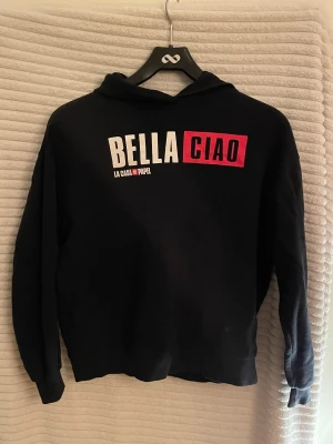 Svart La Casa de Papel hoodie - Svart hoodie med trycket 'BELLA CIAO' i vitt och rött på framsidan och karaktärernas namn i rött på ryggen. Tröjan har huva och långa ärmar. I använt men bra skick. Säljs i befintlig skick. 