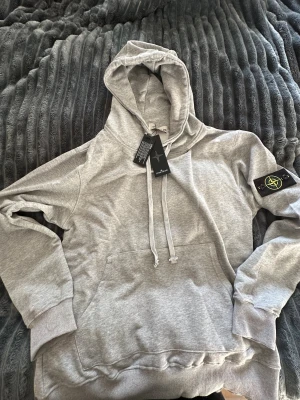 Grå hoodie från Stone Island - Snygg grå hoodie från Stone Island med klassisk patch på vänster ärm. Tröjan har huva med snörning, stor magficka och ribbade muddar. Perfekt för dig som gillar streetwear och vill ha en clean look. Materialet är mjuk bomull för skön komfort.Storlek S&M 170 180