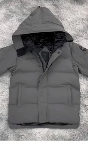 Grå pufferjacka från Canada Goose - Säljer en grå pufferjacka från Canada Goose med huva och snyggt svart märke på ärmen. Jackan har två fickor framtill, dragkedja och knappar, samt ett quiltat foder. Perfekt för kalla dagar och har en clean, modern look.