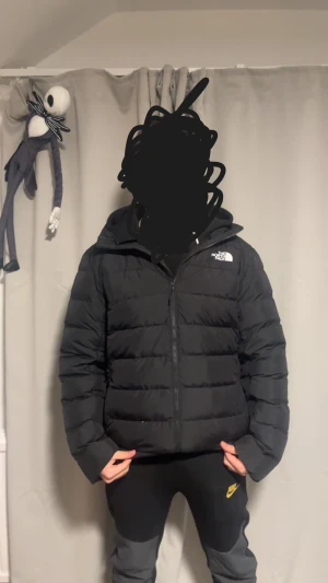 Svart pufferjacka från The North Face - Svart äkta pufferjacka från The North Face med huva och dragkedja framtill. Jackan har ett stilrent, quiltat mönster och logga på bröstet. Perfekt för kalla dagar och har en schysst, lite loose passform. Materialet är syntet och dun för extra värme. Pris diskuteras kan även bytas