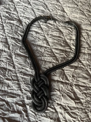 Svart och silverfärgat flätat halsband - Unikt halsband i svart och silver med flätad knutdetalj framtill. Kedjan är grov och har ett justerbart spänne i metall. Perfekt för dig som vill ha ett statement-smycke med cool design och snygg kontrast mellan färgerna.