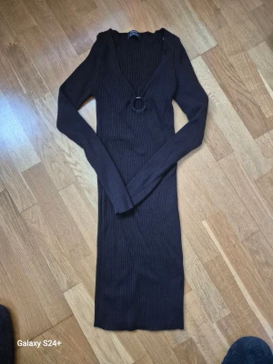 Svart ribbad långklänning Bershka - Snygg klänning från Bershka i svat ribbat material. Klänningen har lång ärm, v-ringad hals med dekorativ ringdetalj framtill och en tight passform som framhäver siluetten. Perfekt för dig som gillar minimalistisk och stilren look.