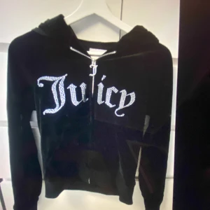 Svart Juicy Couture hoodie med strass - Säljer en äkta Juicy tröja då jag ej använder den och har max haft den på mig 2 gånger.det är en huvtröja från Juicy Couture i mjuk sammet med stor strasslogga 'Juicy ' på bröstet. Tröjan har huva, dragkedja och långa ärmar. Perfekt för dig som gillar den klassiska Y2K-stilen och vill ha något som sticker ut.