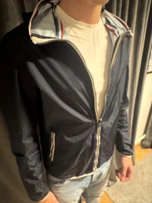 Colmar Windbreaker  - Snygg svart vindjacka från Colmar med huva och dragkedja framtill. Jackan har vita och röda detaljer på insidan av kragen och Colmar-logga på ärmen. Lätt och smidig jacka i syntetmaterial, perfekt för vår och höst. Passformen är normal och jackan har långa ärmar. Pris kan möjligen diskuteras vid snabb köp 