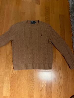Beige kabelstickad tröja Polo Ralph Lauren - Kabelstickad tröja i beige från Polo Ralph Lauren, med klassisk rund hals och röd broderad logga på bröstet. Tillverkad i 100% bomull och har ribbade muddar vid ärmslut och nederkant. Perfekt för dig som gillar stilrena och tidlösa plagg.