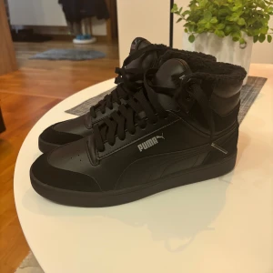 Svarta höga sneakers från Puma - Säljer ett par svarta höga sneakers från Puma i storlek 40. Skorna har snörning, mjukt foder och är tillverkade i skinn och syntet. Perfekta för dig som gillar en clean och stilren look med extra komfort under kallare dagar.