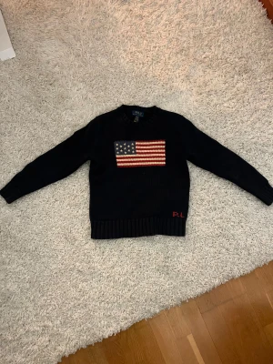 Mörkblå stickad tröja Polo Ralph Lauren - Mörkblå stickad tröja från Polo Ralph Lauren med amerikansk flagga broderad på bröstet och RL-broderi i rött vid nederkanten. Rund hals och långa ärmar, ribbad mudd vid ärmslut och nederkant. 10-12 år