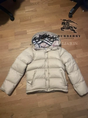Beige Burberry Puffer - Säljer en beige pufferjacka från Burberry med klassiskt rutigt foder i huvan. Jackan har dragkedja och knappar framtill, två fickor med dragkedja och en mysig, vadderad känsla. Perfekt för kalla dagar och riktigt snygg med den ikoniska Burberry-looken.