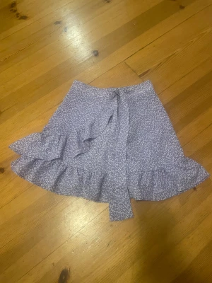 Ljuslila omlottkjol med volang - Supersöt ljuslila kortkjol från newyorker i omlottmodell med volangkant. Kjolen har ett vitt småmönstrat tryck och knytband i midjan. Perfekt för dig som gillar feminina detaljer och vill ha en kjol som sticker ut lite extra.