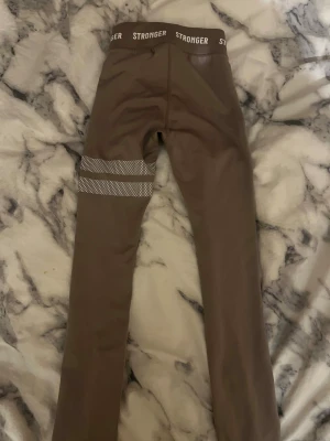 Beige leggings från Stronger XXS - Snygga beiga leggings från Stronger i storlek XXS. De har ett elastiskt midjeband med logga och coola vita prickiga detaljer på ena benet. Materialet är mjukt och stretchigt polyester, perfekt för träning eller chill. Passformen är tight och midjan är hög.