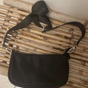 Svart axelväska från Lindex - Svart axelväska från Lindex med stilren, rundad form och bred justerbar axelrem. Väskan är tillverkad i syntetmaterial och har en silverfärgad dragkedja upptill. Perfekt storlek för mobil, plånbok och småsaker. Enkel och clean design.