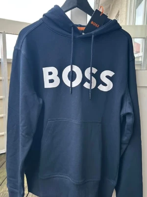 Helt ny Marinblå hoodie från BOSS med tryck - Snygg marinblå hoodie från BOSS med stort vitt BOSS-tryck på bröstet. Tröjan har huva med snörning, känguruficka och långa ärmar. Tillverkad i mjuk bomull och polyesterblandning för skön komfort. Perfekt för dig som gillar stilrena och trendiga plagg.