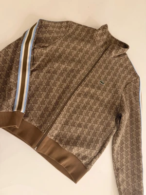 Brun mönstrad trackjacka från Lacoste - Snygg brun trackjacka från Lacoste med geometriskt mönster och klassisk krokodillogo på bröstet. Jackan har ribbade muddar, dragkedja framtill och coola ränder i blått, vitt och brunt längs ärmarna. Perfekt för dig som gillar sportig och trendig stil.
