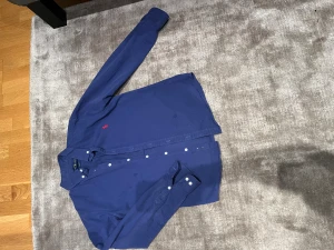 Blå skjorta från Ralph Lauren - För nu läget har den fläckar men du som köpare får den självklart nytvättad! Annars är de en Snygg blå skjorta från Ralph Lauren med klassisk röd broderad logga på bröstet. Skjortan har vita knappar framtill och är långärmad med krage. Tillverkad i mjuk bomull som känns skön mot huden. Perfekt för dig som gillar stilrena och tidlösa plagg. Pris kan diskuteras! 