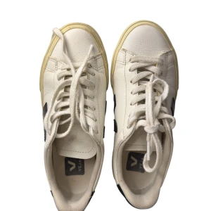 Veja vita sneakers med svart logga - Snygga vita sneakers från Veja med svart V-logga på sidan. Skorna har rund tå, platt sula och klassisk snörning. Tillverkade i skinn med beige detaljer på sulan.