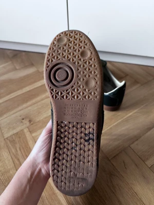 Maison Margiela oliv gröna sneakers - Maison Margiela sneakers i oliv grönt med mörkgrå mockadetaljer och vita snören. Snygg retrostil med bruna gummisulor och rund tå. Ovandelen är en mix av skinn och mocka, vilket ger en cool kontrast. Perfekt för dig som gillar stilrena och unika sneakers. Skriv gärna vid fler frågor! 