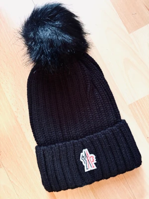 Svart ribbstickad mössa från Moncler! - Svart ribbstickad mössa med avtagbar tofs i Onesize från Moncler. Oanvänd.