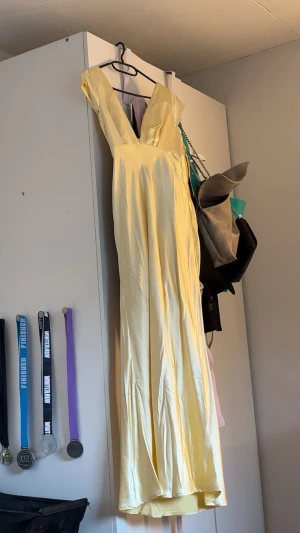 Ljusgul långklänning med v-ringning - Säljer en elegant ljusgul långklänning med djup v-ringning och korta ärmar. Klänningen har en figurnära passform upptill och faller snyggt längs kroppen. Materialet är glansigt och känns lyxigt, perfekt för student eller bal.