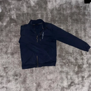 Mörkblå jacka från Sail Racing - Mörkblå jacka från Sail Racing med hel dragkedja och en extra dragkedja på bröstfickan. Jackan har ståkrage och långärmade ärmar. Materialet är mjukt och känns som polyester, perfekt för kyliga dagar. Snygg logga broderad på bröstet. Pris kan diskuteras!