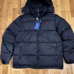 Svart pufferjacka från Polo Ralph Lauren - Svart pufferjacka från Polo Ralph Lauren med röd broderad logga på bröstet. Jackan har huva, två sidofickor med dragkedja och hel dragkedja framtill. Materialet är polyester och jackan har en klassisk, normal passform som är perfekt för kalla dagar.