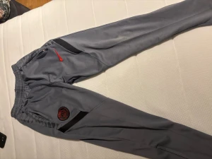 Grå PSG mjukisbyxor från Nike - Snygga grå mjukisbyxor från Nike med Paris Saint-Germain-logga på benet och röd Nike-swoosh. Byxorna har svarta detaljer längs sidorna och elastisk midja för skön passform. Perfekta för chill eller träning.