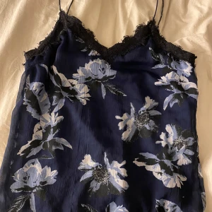 Blommig spetslinne - Säljer ett vackert blommigt linne i blått med tunna axelband och spetsdetaljer vid halsringningen. Perfekt för en somrig look. Blommönstret går i ljusblå och vita nyanser.
