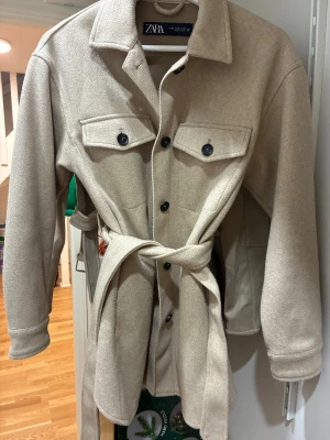 Beige overshirt jacka från Zara - Snygg beige overshirt jacka från Zara med klassisk krage, stora bröstfickor med knappar och bälte i midjan. Jackan har långa ärmar med knappdetalj vid mudden och är tillverkad i ett mjukt ull-liknande material. Perfekt för lager-på-lager och chill stil.