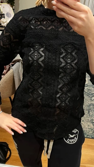 Svart spetsblus med mönster - Säljer en svart blus med lång ärm och transparenta partier i spets. Blusen har geometriskt broderat mönster och hög hals. Materialet är lätt och ger en cool vibe med sina detaljerade spetsar och mönster. Perfekt för dig som gillar att sticka ut med stil.