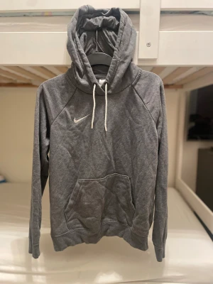 Grå hoodie från Nike med huva - Grå hoodie från Nike med vit logga på bröstet och vita snören i huvan. Tröjan har en stor magficka och är långärmad. Mjuk och skön bomullsmix som passar perfekt när du vill chilla eller dra till gymmet. Klassisk och enkel stil med normal passform. 